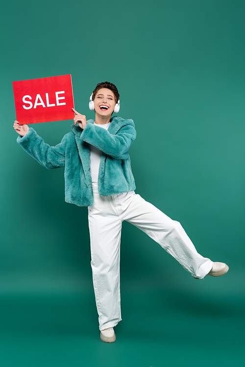 excited woman in faux fur jacket, white pants and headphones holding card with sale lettering on green