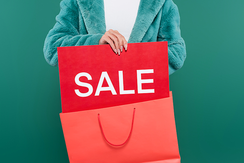 cropped view of woman in faux fur jacket holding pink shopping bag and credit card with sale lettering isolated on green
