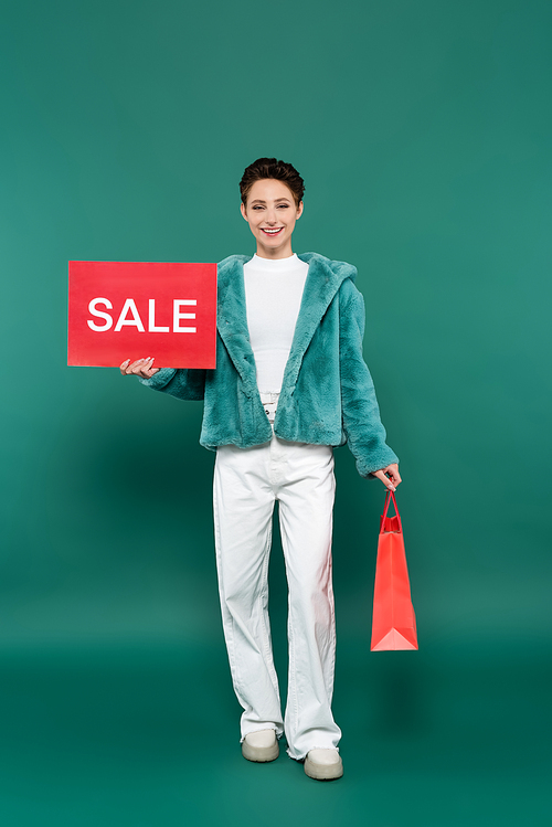 happy woman in faux fur jacket and white pants holding shopping bag and red card with sale lettering on green