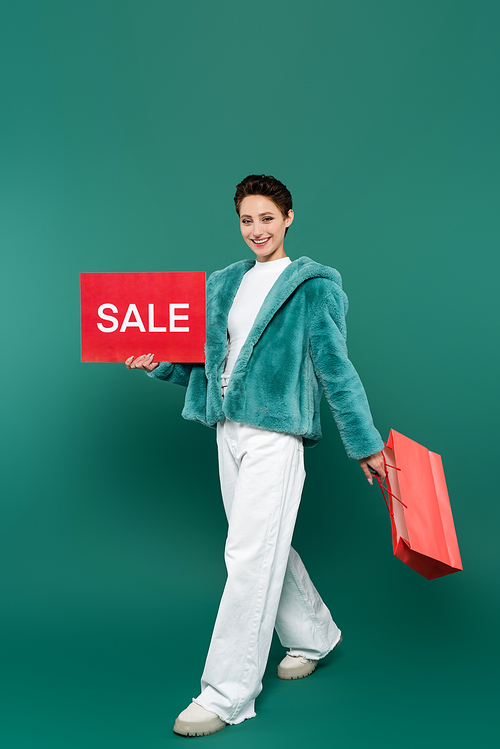 cheerful woman in white pants and faux fur jacket walking with sale card and shopping bag on green