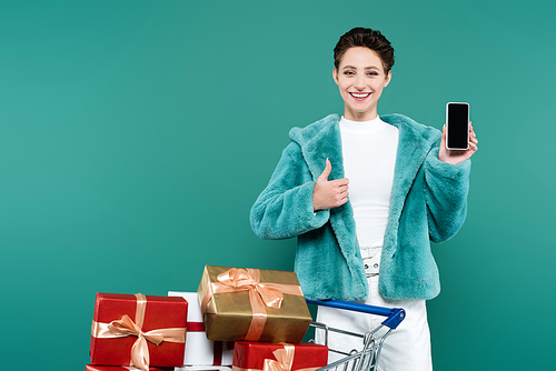 smiling woman showing thumb up while holding smartphone with blank screen near shopping cart with gifts isolated on green