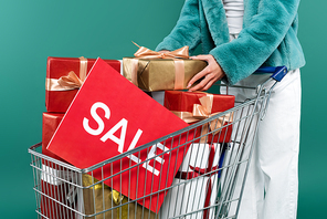 partial view of trendy woman near shopping cart with gift boxes and card with sale lettering isolated on green