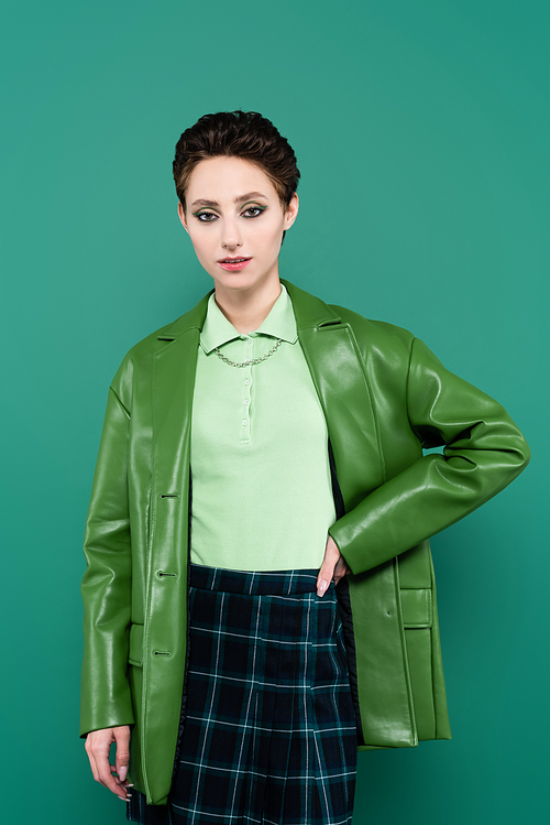 young woman in leather jacket and plaid skirt posing with hand on hip isolated on green