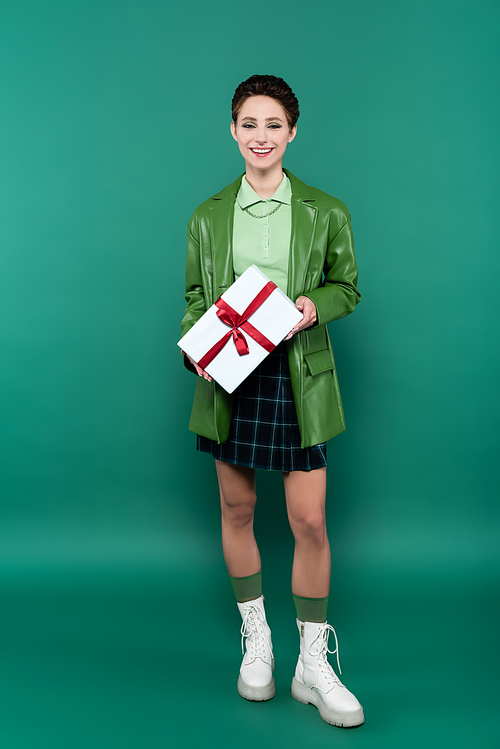 full length view of happy woman in leather jacket, checkered skirt and white boots posing with gift box on green