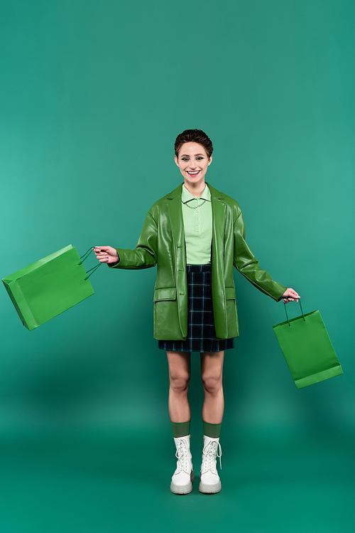 full length view of smiling woman in leather jacket, plaid skirt and white boots posing with shopping bags on green