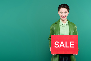 pleased woman in leather jacket holding red card with sale lettering isolated on green