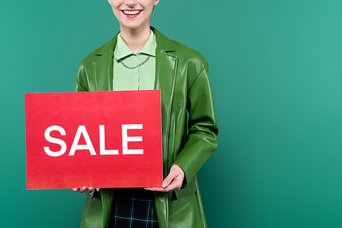 cropped view of woman in stylish jacket holding red card with sale lettering isolated on green