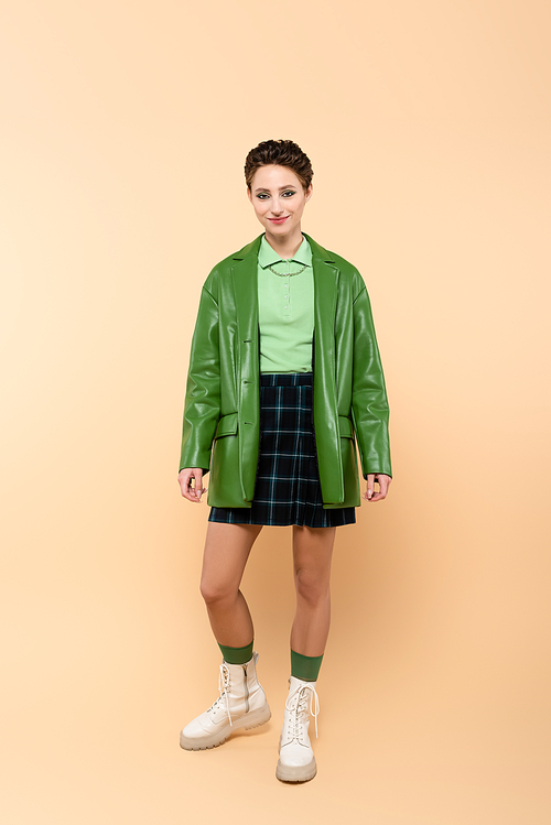 full length view of positive woman in green jacket, plaid skirt and white lace-up boots on beige