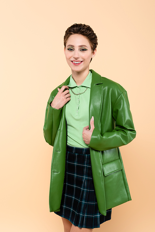 brunette woman in green leather jacket and plaid skirt smiling at camera isolated on beige
