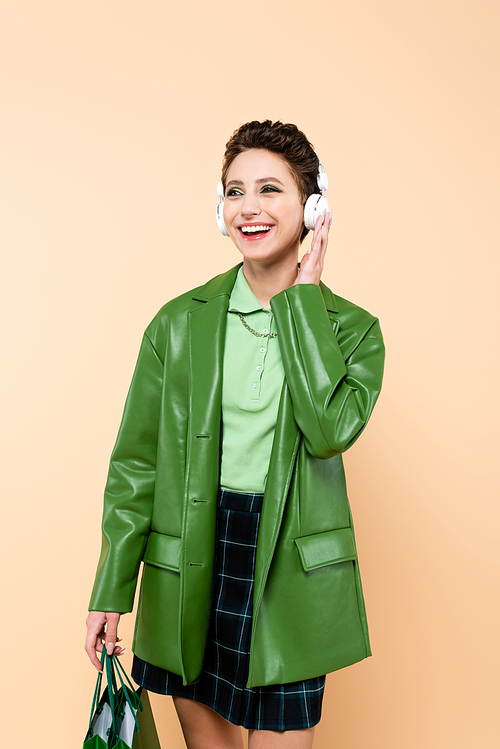 joyful and fashionable woman with shopping bags adjusting headphones isolated on beige