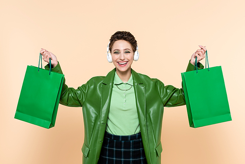 excited woman in headphones and green jacket holding purchases isolated on beige