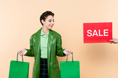 man holding red card with sale lettering near trendy woman with green shopping bags on beige