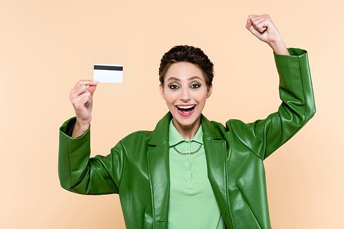 excited woman in green leather jacket showing success gesture while holding credit card isolated on beige
