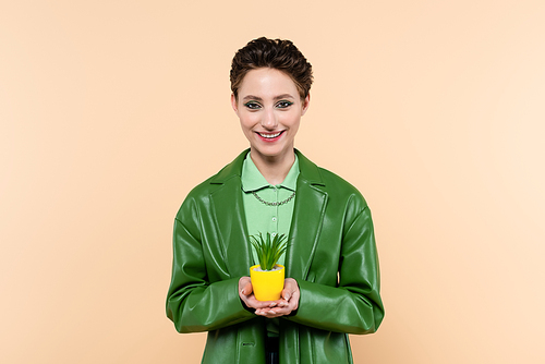happy woman in green jacket holding plant in yellow flowerpot isolated on beige
