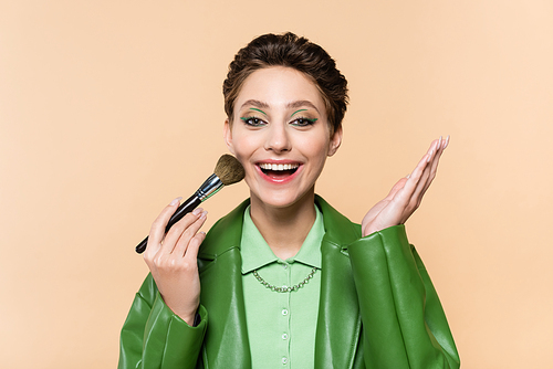 excited woman applying face powder with cosmetic brush and pointing with hand isolated on beige