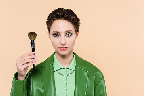 brunette woman in green leather jacket holding cosmetic brush isolated on beige