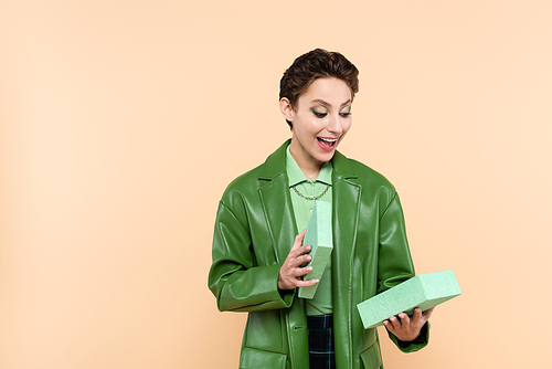amazed woman in green leather jacket looking in gift box isolated on beige