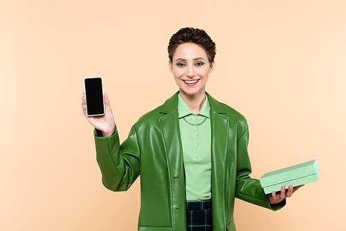 cheerful brunette woman with gift box showing smartphone with blank screen isolated on beige