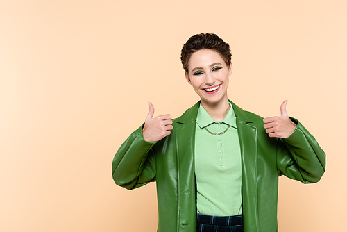 joyful woman in green stylish jacket showing thumbs up isolated on beige