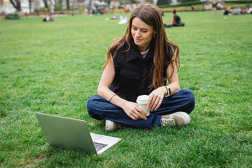 cheerful young woman holding paper cup and looking at laptop while sitting on lawn