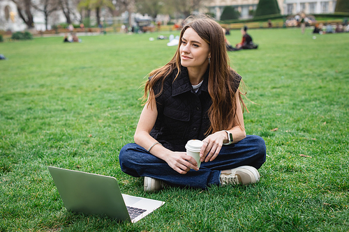 cheerful young woman holding paper cup near laptop while sitting on lawn