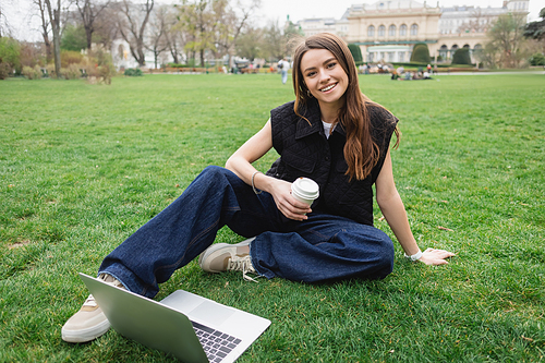 smiling young woman holding paper cup near laptop while sitting on lawn