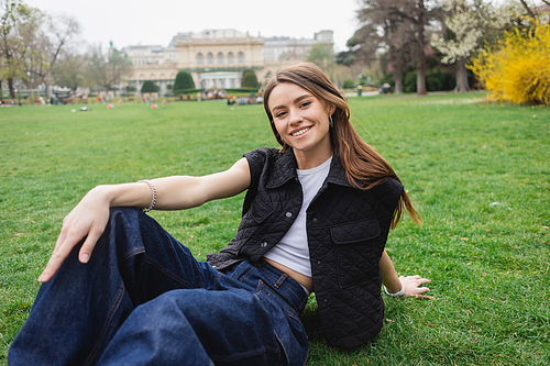 joyful young woman in sleeveless jacket sitting on lawn