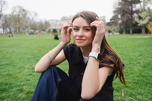 smiling young woman in sleeveless jacket adjusting hair while sitting on lawn