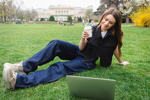 happy young woman in sleeveless jacket holding paper cup and resting on lawn near laptop