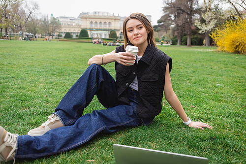 thoughtful young woman in sleeveless jacket holding paper cup and resting on lawn near laptop