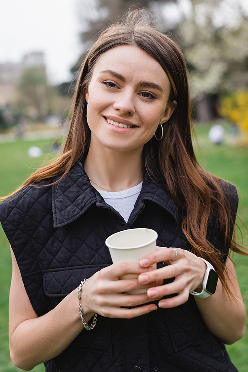 young cheerful woman in sleeveless jacket holding coffee to go in green park