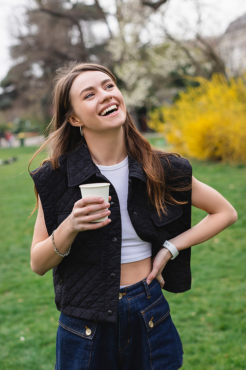 young joyful woman in sleeveless jacket holding coffee to go in green park