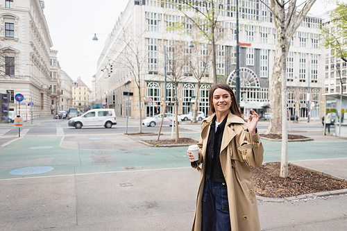 happy young woman in stylish trench coat walking with coffee to go and waving hand in european city