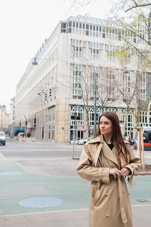 young woman adjusting belt on stylish trench coat on street of european city