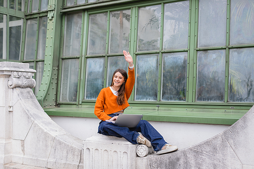 happy young woman waving hand while using laptop on rooftop of building in vienna