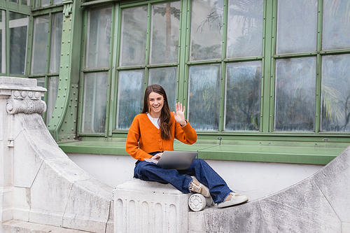 cheerful young woman waving hand while using laptop on rooftop of building