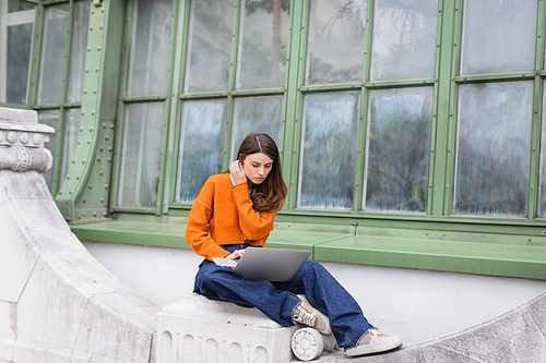 young woman in jeans and orange cardigan using laptop on rooftop of building