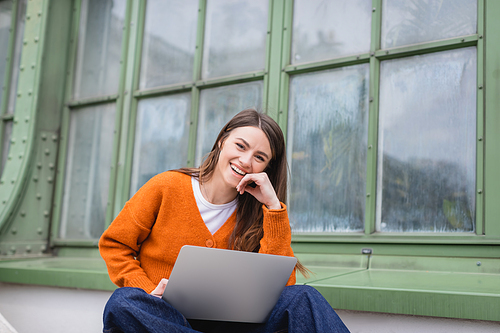 happy young woman in jeans and orange cardigan using laptop on rooftop of building