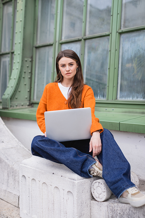 young woman in jeans and orange cardigan sitting with laptop on rooftop of building