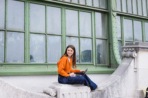 happy young freelancer in jeans and orange cardigan sitting with laptop on rooftop of building