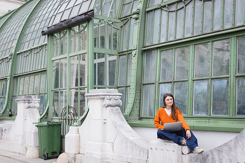 smiling young freelancer in jeans and orange cardigan sitting with laptop on rooftop of building