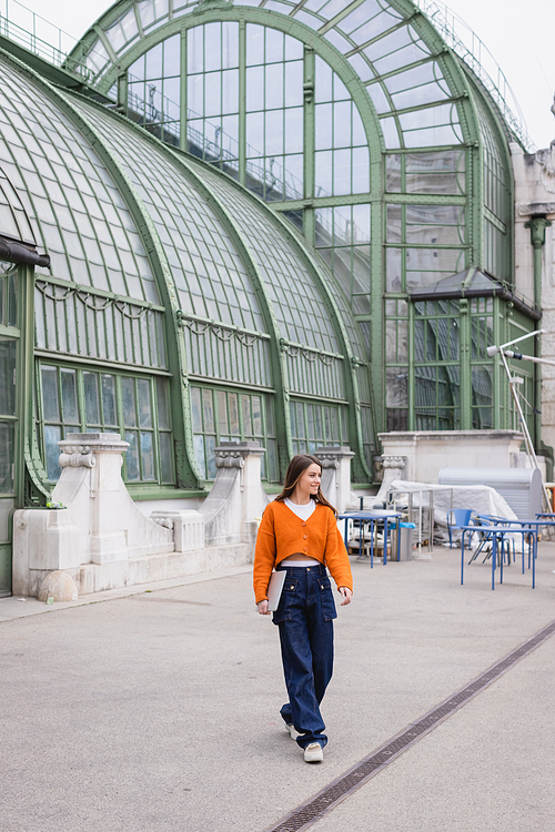 happy young woman in jeans and orange cardigan walking with laptop on rooftop of building