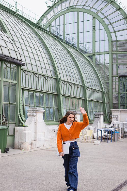 happy young woman waving hand and walking with laptop on rooftop of building