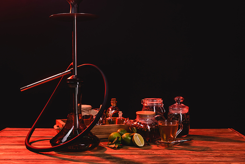 Tea, fresh limes and hookah on wooden table isolated on black with red light