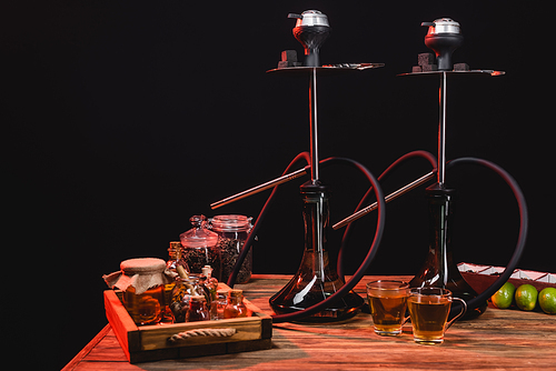 Tea cups near hookahs and limes on wooden surface isolated on black