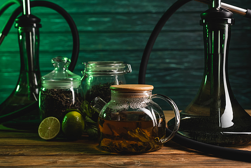 Teapot near blurred limes and hookahs on wooden table on wooden background