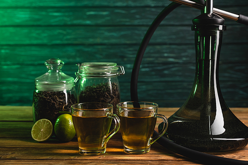 Cups of tea, limes and hookah on wooden surface on wooden background