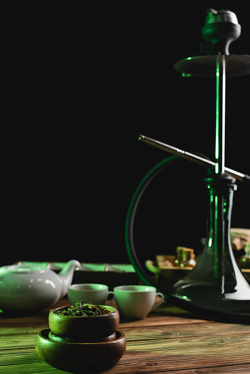 Dry tea near blurred cups and hookah on wooden surface isolated on black