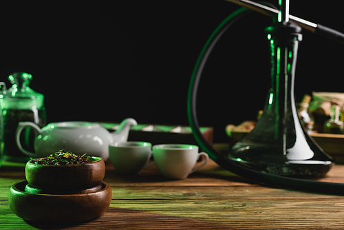 Dry tea near blurred hookah and cups on wooden table isolated on black