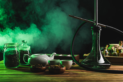 Hookah near dry tea and teapot on wooden surface on black background with smoke